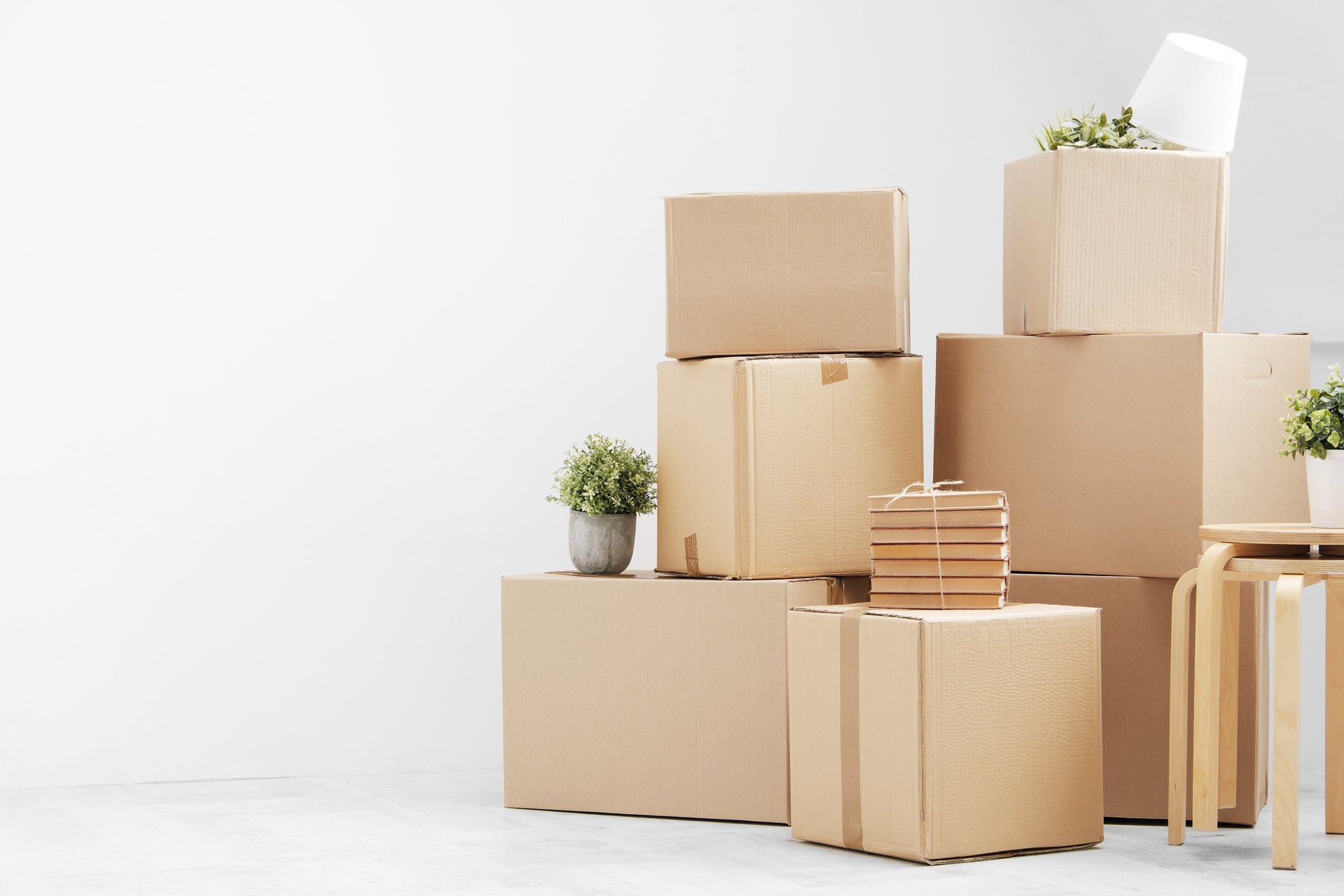 Moving to a new home. Belongings in cardboard boxes, books and green plants in pots stand on the gray floor against the background of a white wall.